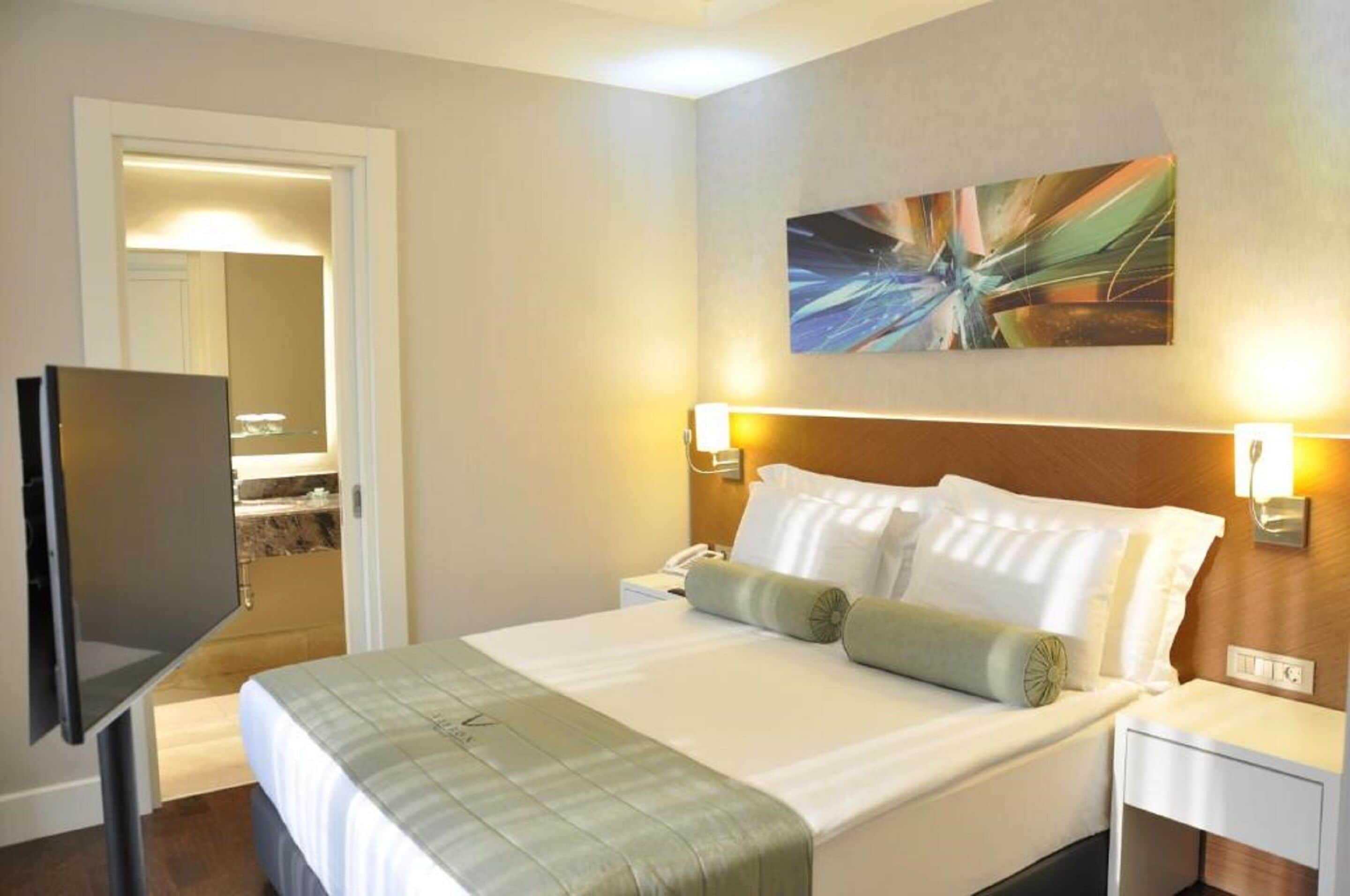 Habitación de hotel con cama, lámparas de noche, televisor de pantalla plana, un cuadro en la pared y una puerta que da al baño.