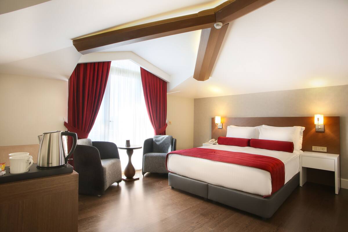 Una habitación de hotel con una cama grande, un escritorio con hervidor, dos sillones y una mesita.