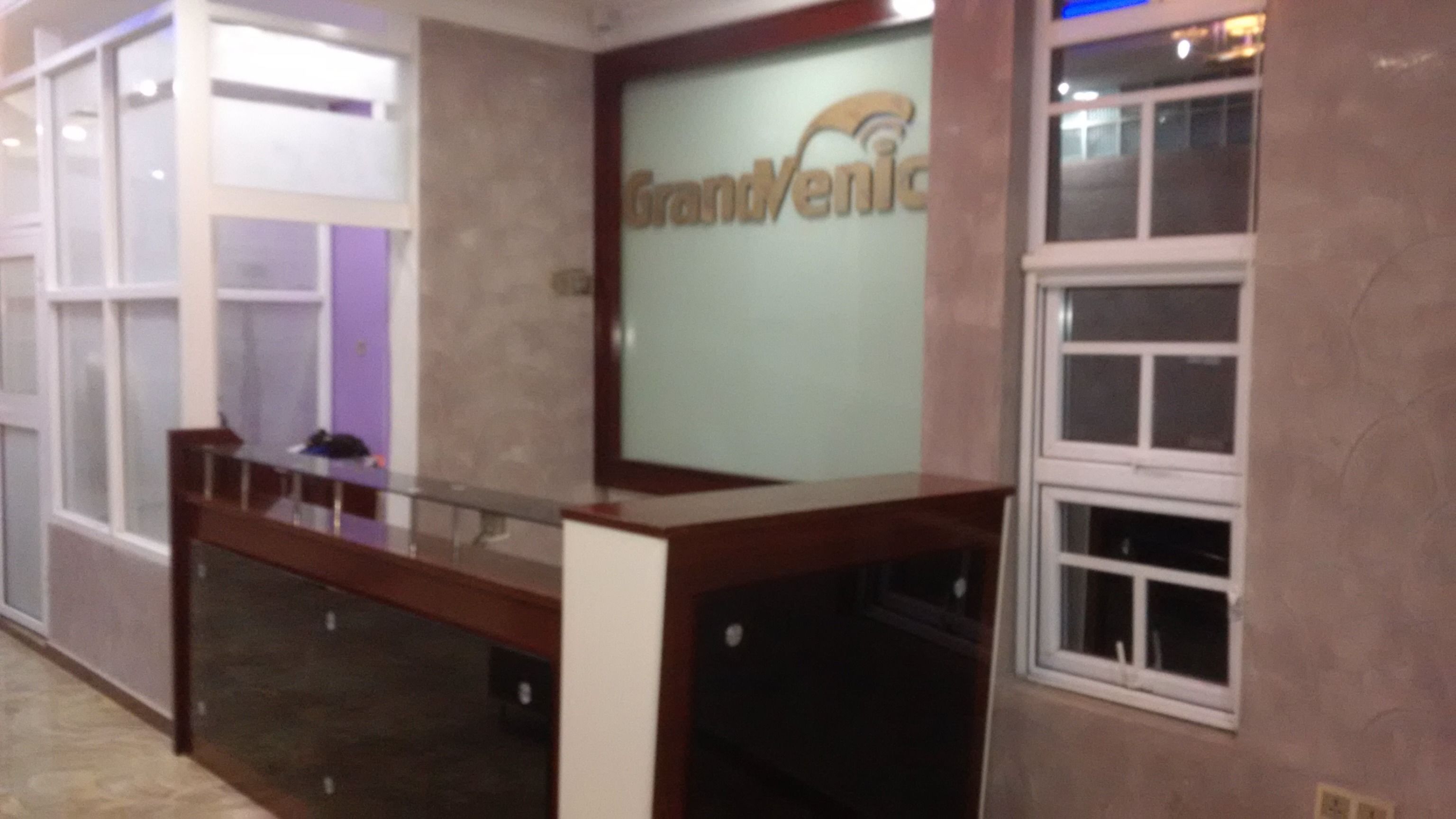 A reception desk with a glass partition, a sign reading "GrandVenic", and a window with blinds.
