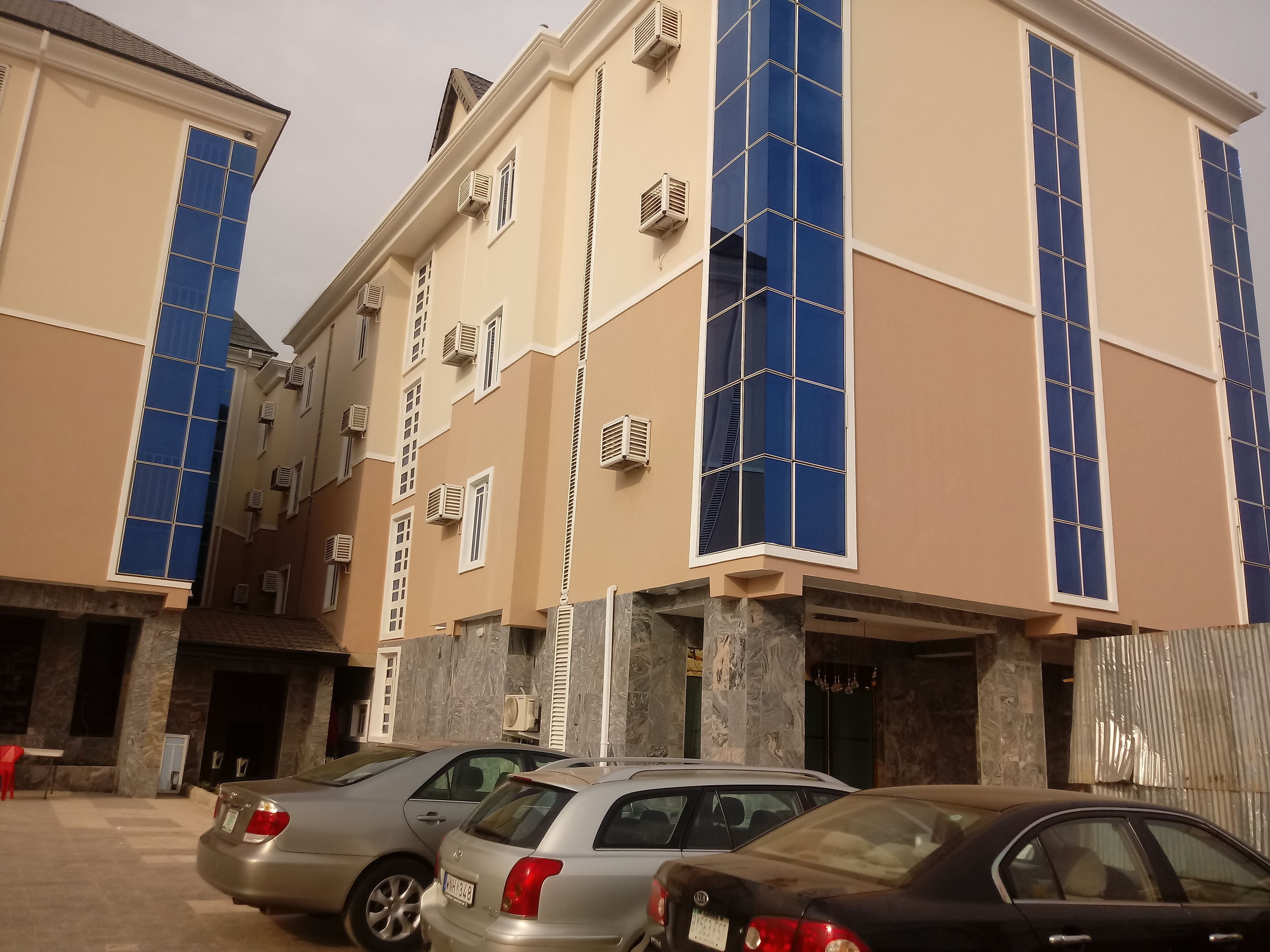 A multi-story building with large windows and air conditioning units. There are several parked cars in front.