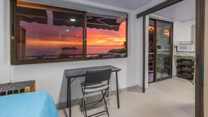 Une pièce avec une grande fenêtre donnant sur un coucher de soleil, un bureau avec une chaise et une petite table recouverte d’une nappe bleue.