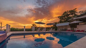 Une piscine entourée d’une clôture et d’arbres, avec des chaises longues et des parasols, au moment du coucher du soleil.
