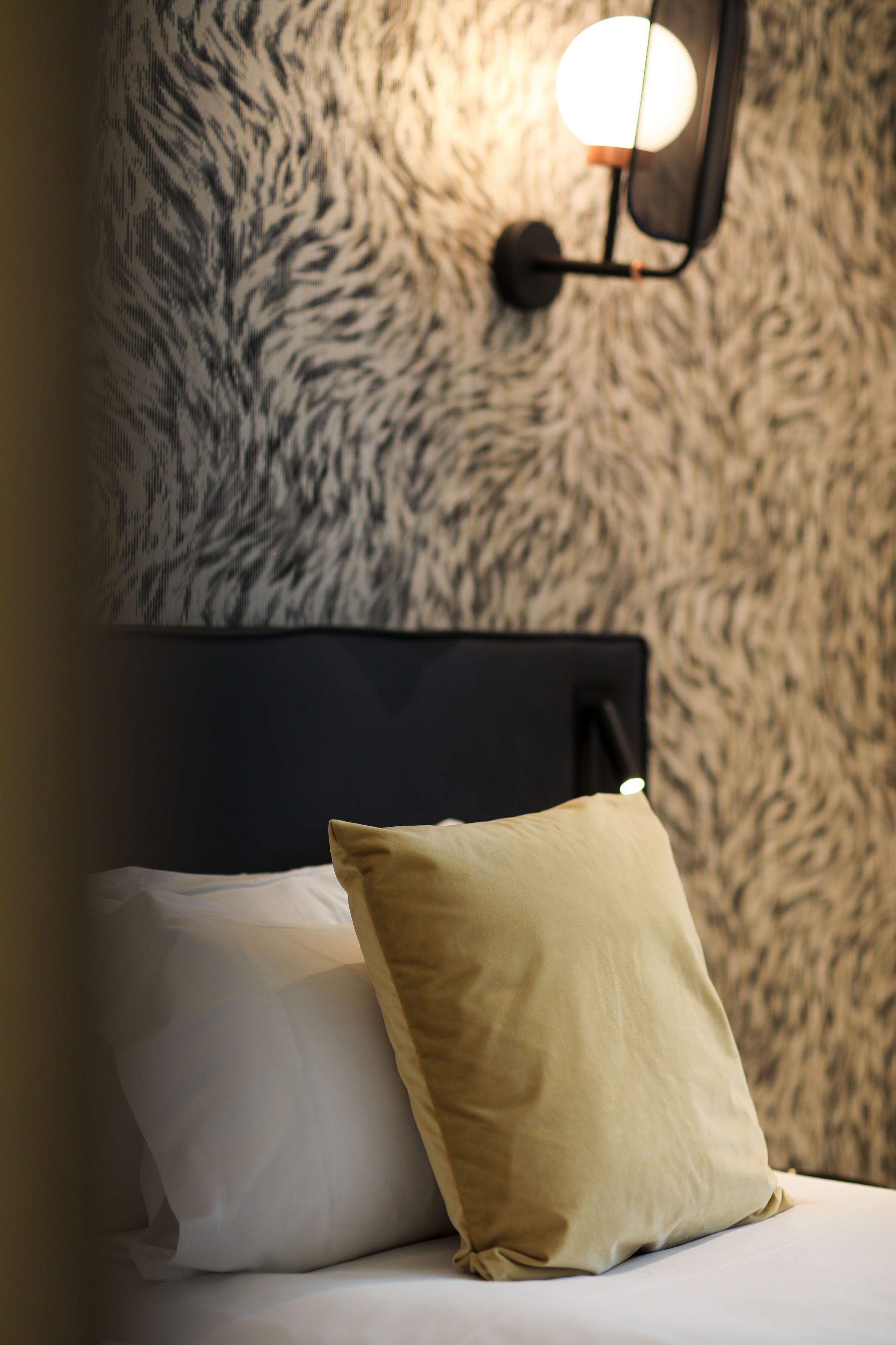 A bed with white and beige pillows, a black headboard, and a textured wall with a wall-mounted light fixture.