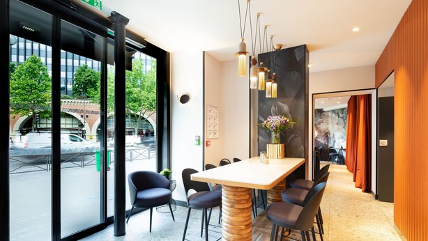 Un moderno vestíbulo de hotel con una mesa grande y sillas, un mostrador de recepción y ventanas amplias que ofrecen vistas a la calle y a los árboles.