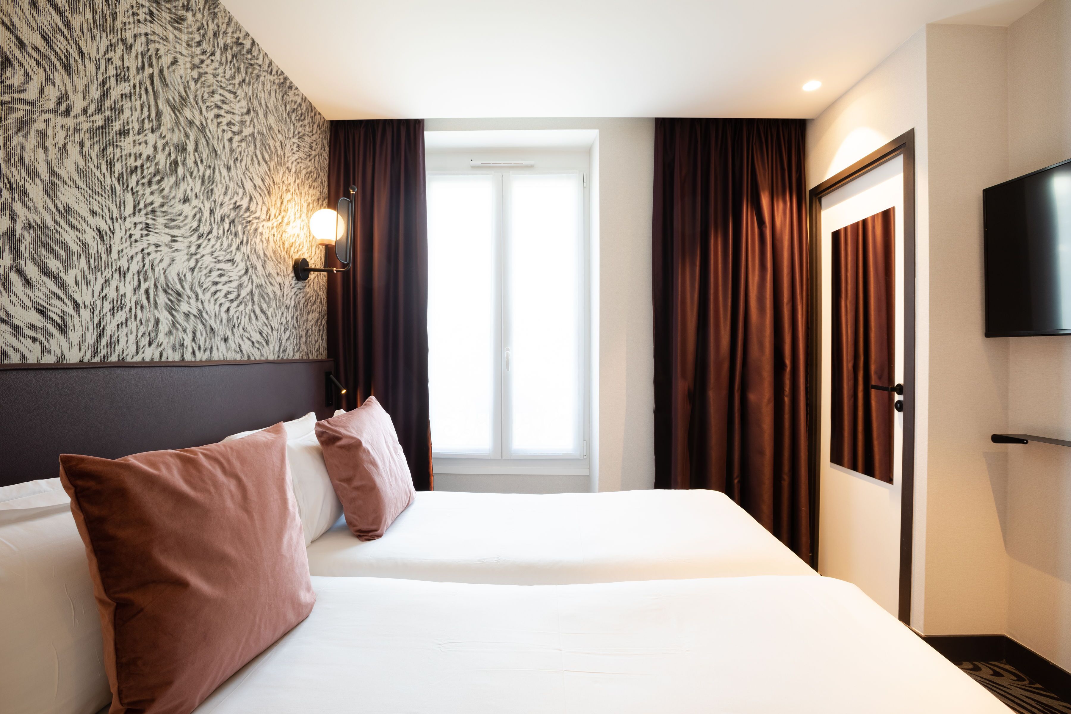A hotel room with a large bed, a wall-mounted TV, and a window with curtains.