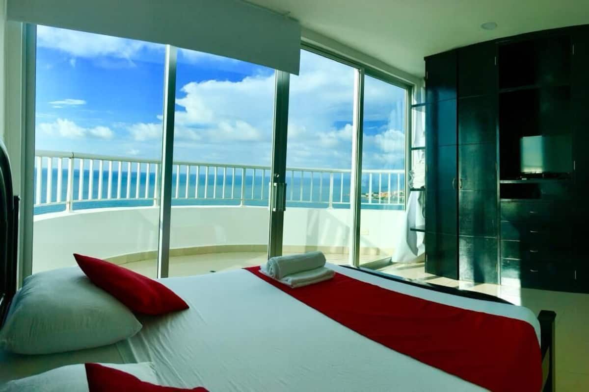 A bedroom with a large bed, a balcony with a view of the sea, and a dark-colored wardrobe.