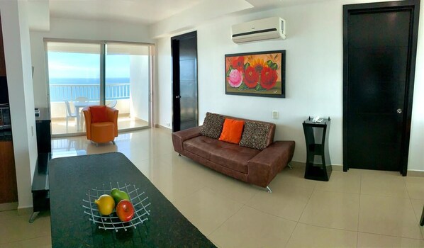 A living room with a sofa, a dining table with fruit, a painting, and a sliding glass door to a balcony. - Apartamentos frente al mar Cartagena - Edificio Palmetto (Cartagena)