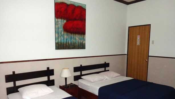 A hotel room with two single beds, a nightstand with a lamp, a wooden door, and a painting of red flowers on the wall. - Hotel Santamaria (Alajuela)