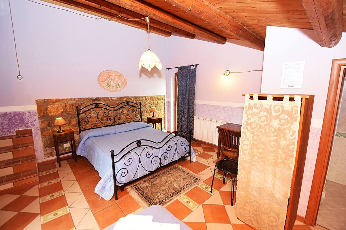 Un dormitorio con cama, mesita de noche, lámpara, silla de madera y cortina estampada.