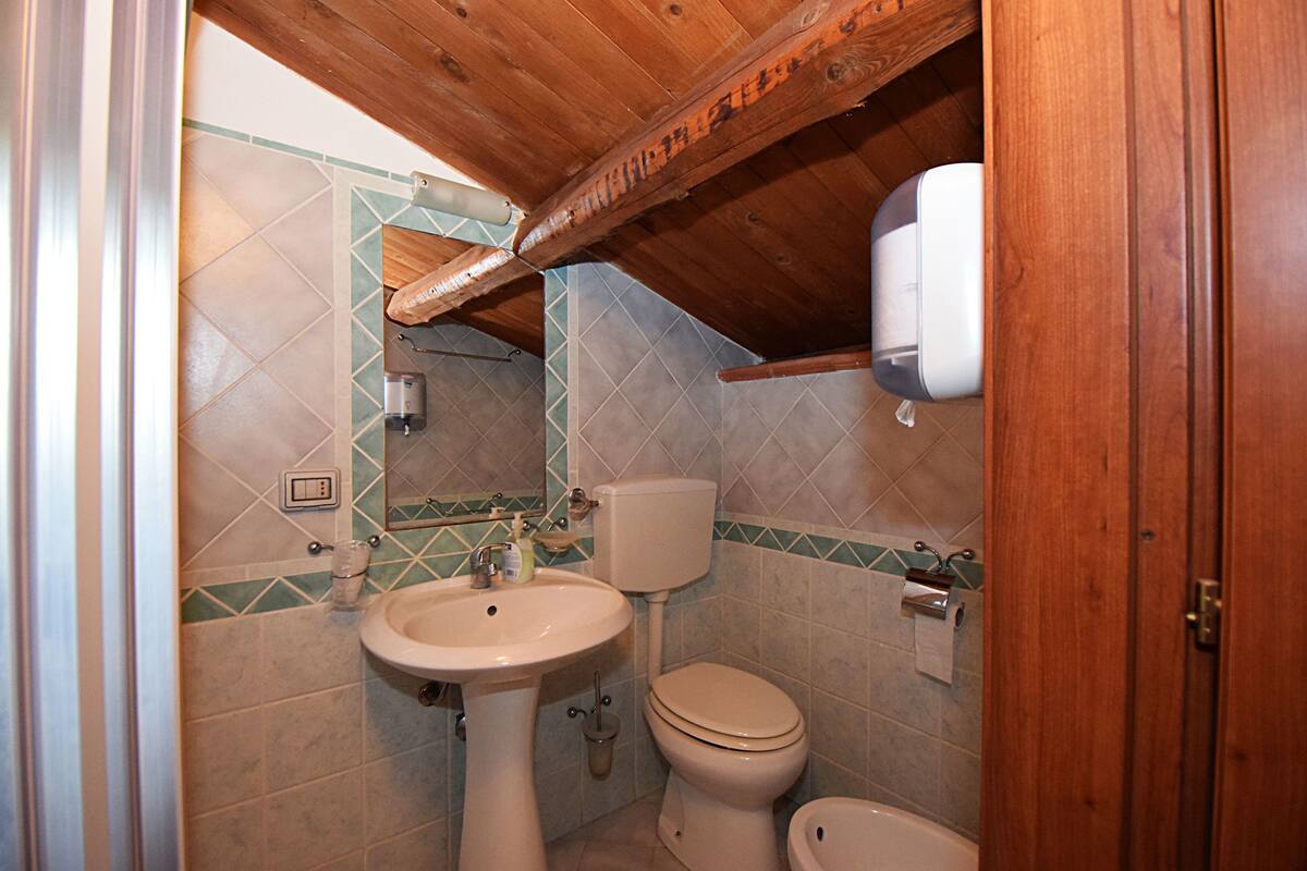 Un baño con lavamanos pedestal, inodoro y bidé, con azulejos verde y beige y vigas de madera en el techo.