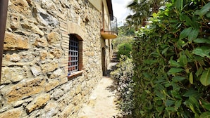 Un étroit sentier de pierre, bordé d’un bâtiment en pierre et d’une végétation luxuriante.