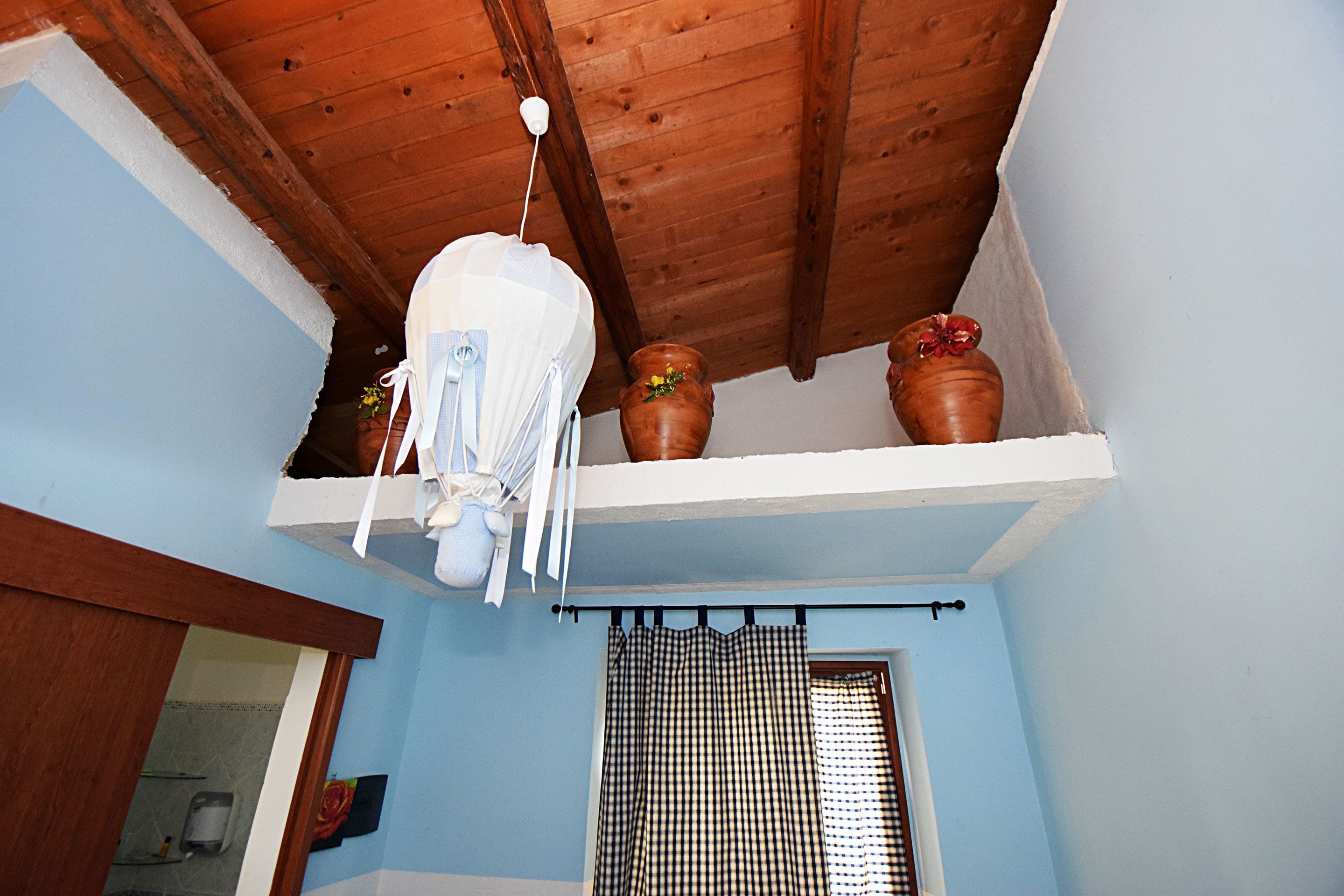 A room with a wooden ceiling, a white shelf with terracotta pots, and a checkered curtain.