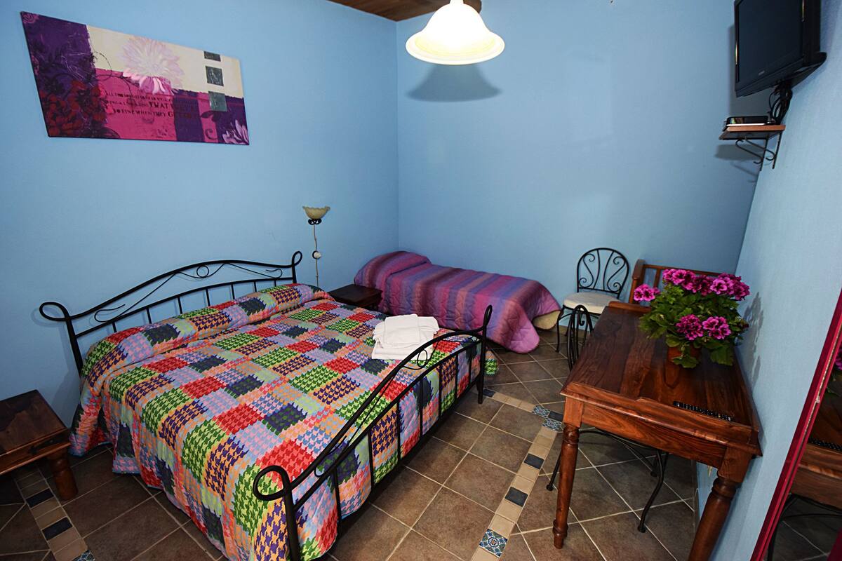 Un dormitorio con cama, mesita de noche, mesa de madera con un florero y televisor montado en la pared.