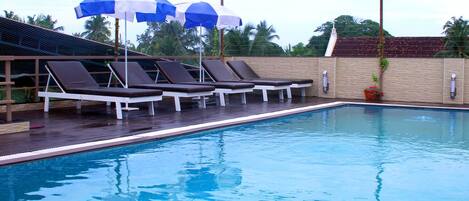 A swimming pool with lounge chairs and umbrellas, surrounded by a wooden deck and a brick wall.