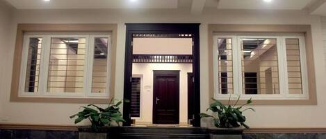 A building entrance with a dark wooden door, two large windows with white frames, and two potted plants on either side.