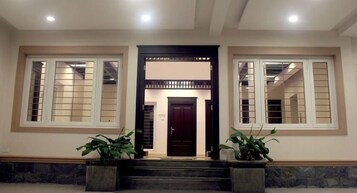 A building entrance with a dark wooden door, two large windows with white frames, and two potted plants on either side.