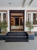 A building entrance with a dark wooden door, steps leading up to a landing, and potted plants on either side.