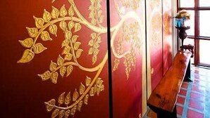 A hallway with a red wall featuring a golden tree design, a wooden bench, and a window with natural light. - Huenchandee (Chiang Rai)