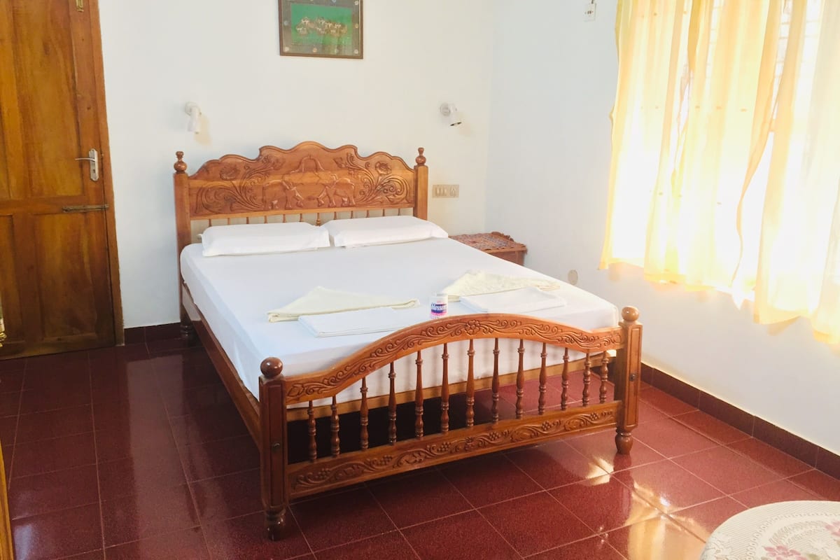 A bedroom with a wooden bed, white linens, a nightstand, a wall-mounted picture, and a window with curtains.