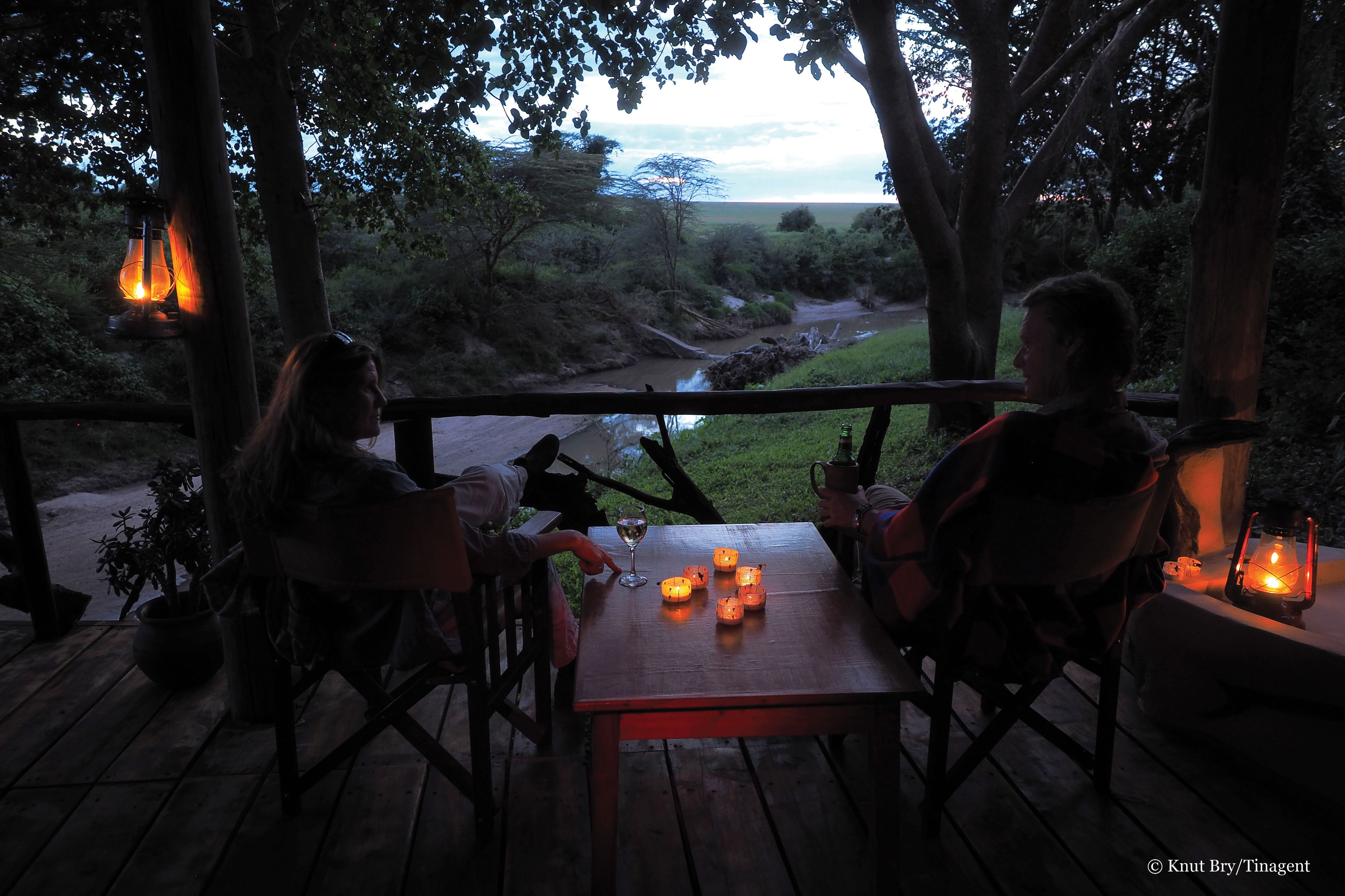 An outdoor wooden deck with a table and chairs, surrounded by trees and a view of a river.