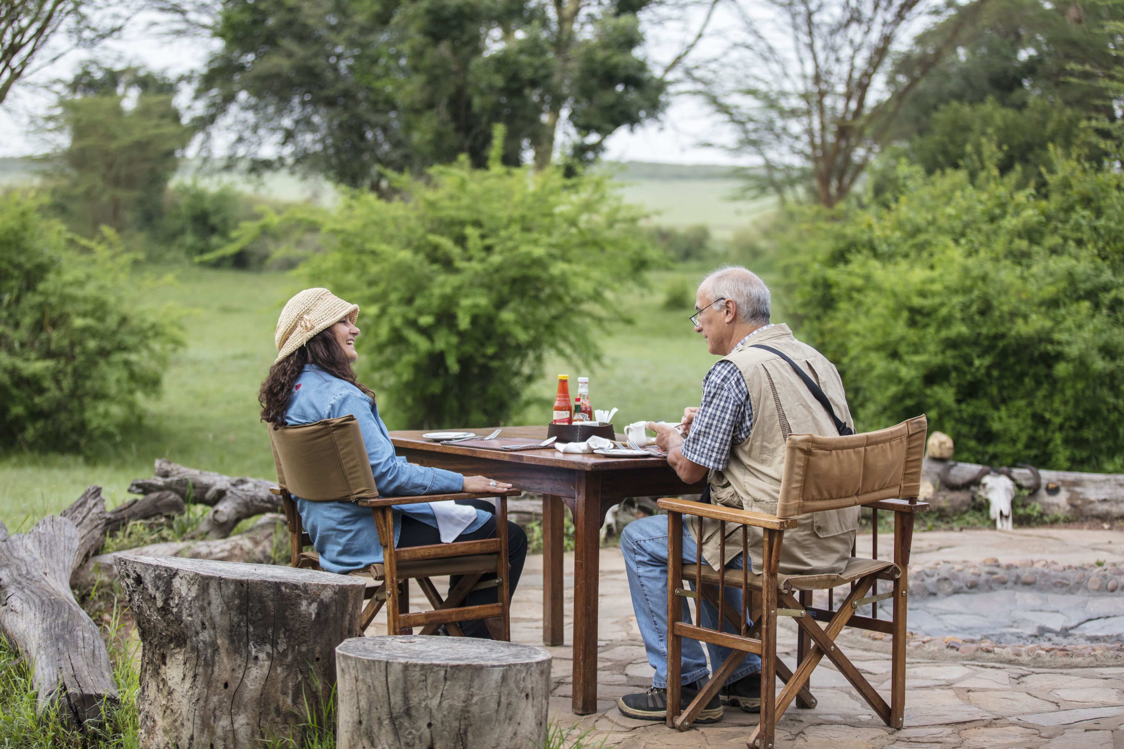 Two people seated at an outdoor wooden table with a view of a grassy field and trees in the background.
