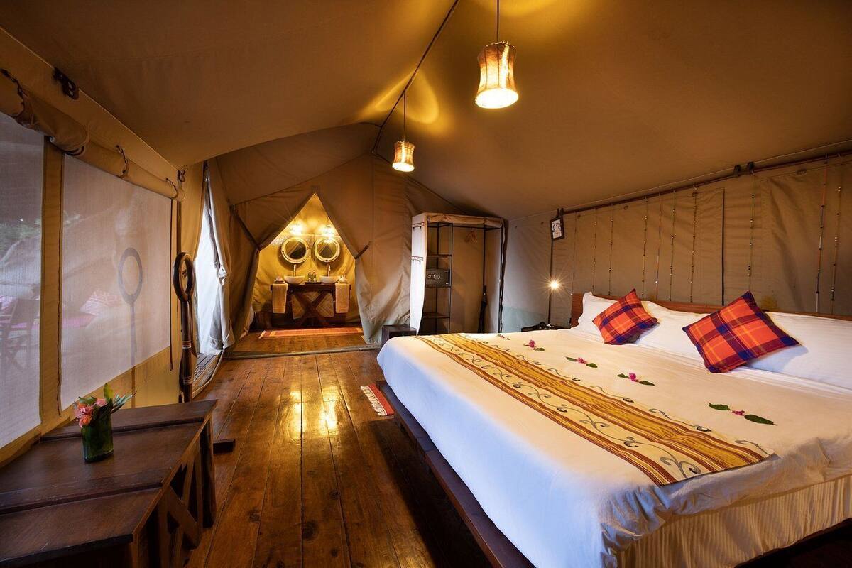 A cozy tent-style room with a wooden floor, a bed with a patterned bedspread, two bedside lamps, and a small table with a vase of flowers.