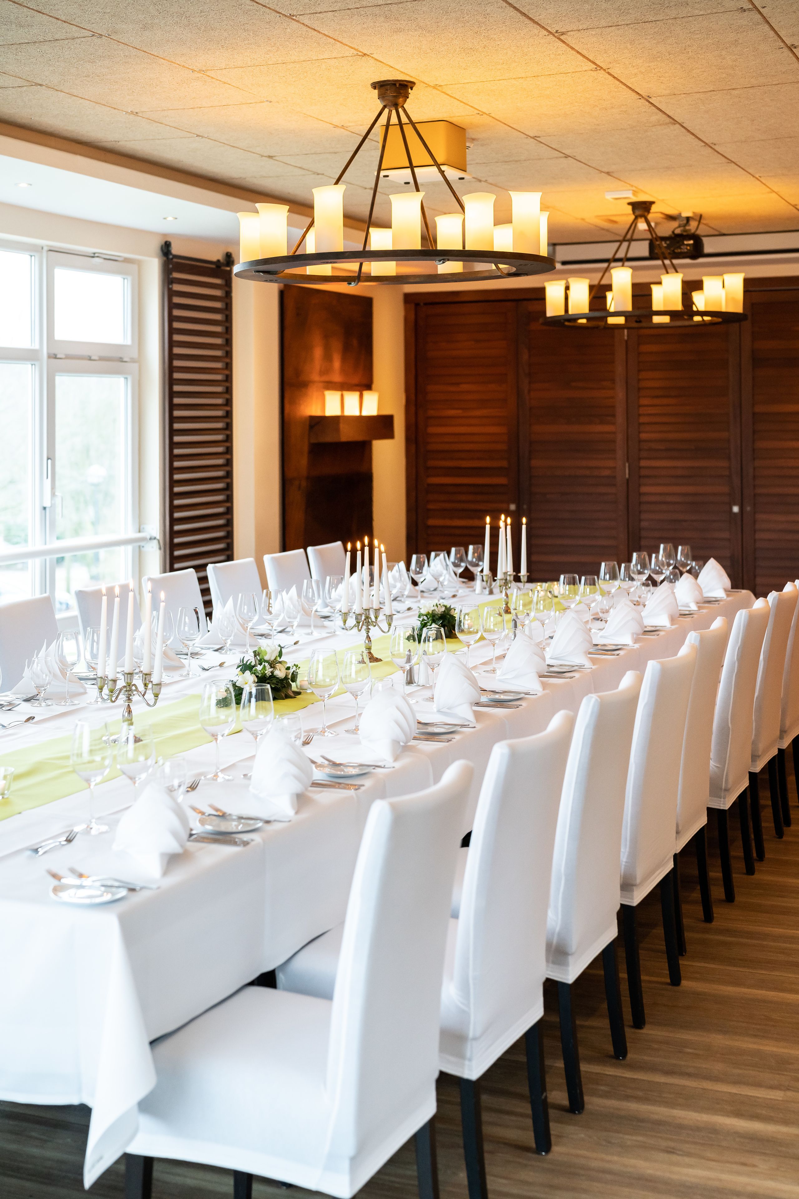 Una larga mesa de comedor cubierta con manteles blancos, velas y sillas, en una sala con paneles de madera y un candelabro.