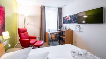 Ein Hotelzimmer mit einem Bett, einem Schreibtisch, einem roten Sessel, einem Fernseher und einem Bild an der Wand.