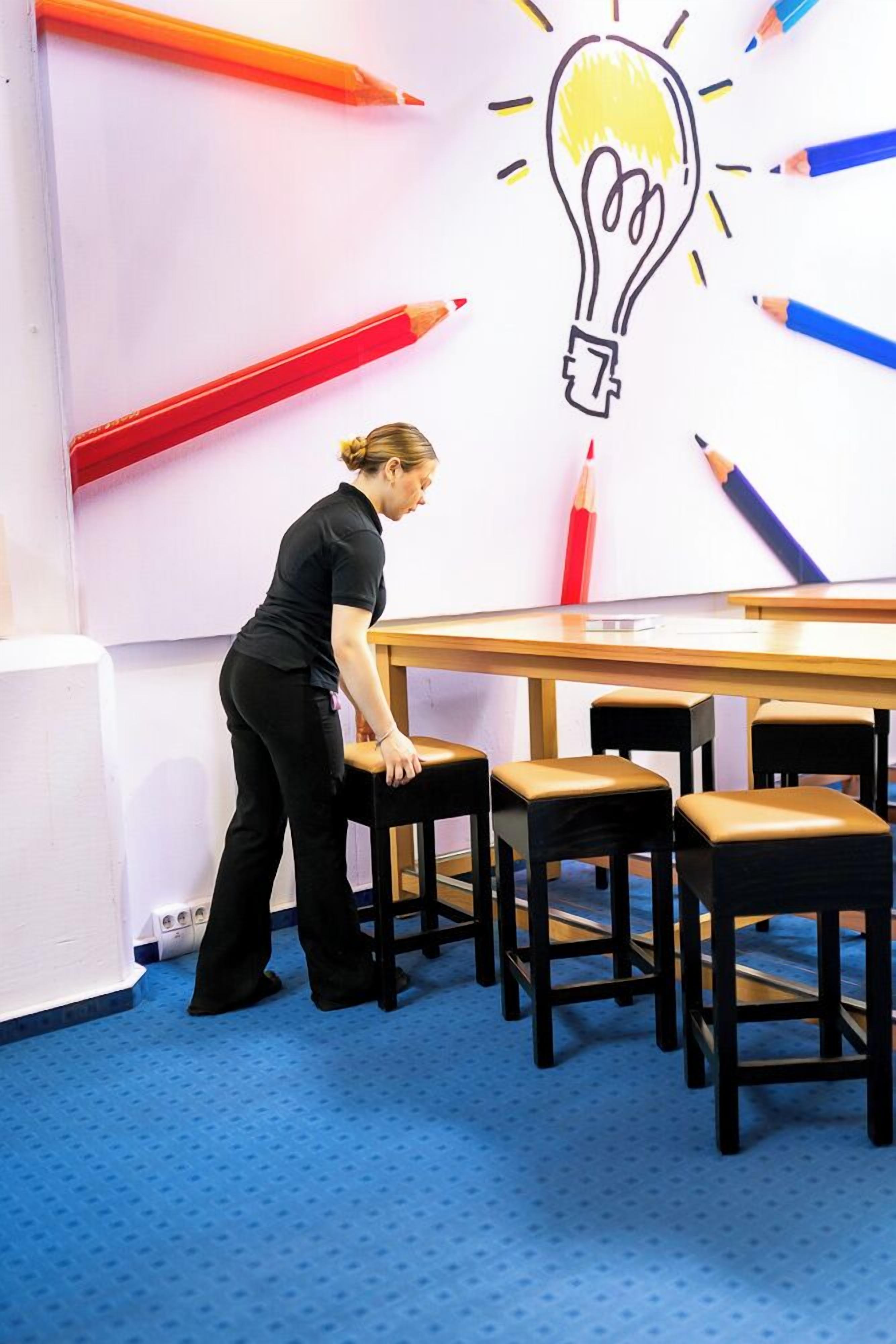 A person standing by a table with a light bulb drawing on the wall, surrounded by chairs.