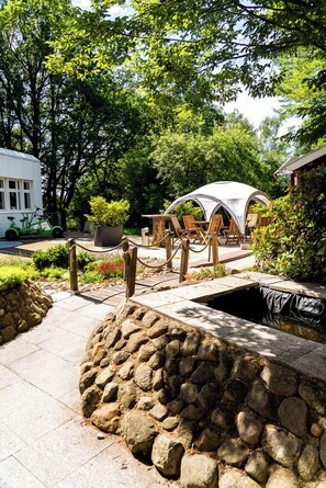 A stone pathway leading to a covered seating area with chairs and a table, surrounded by greenery and trees. - ANDERS Hotel Walsrode (Walsrode)