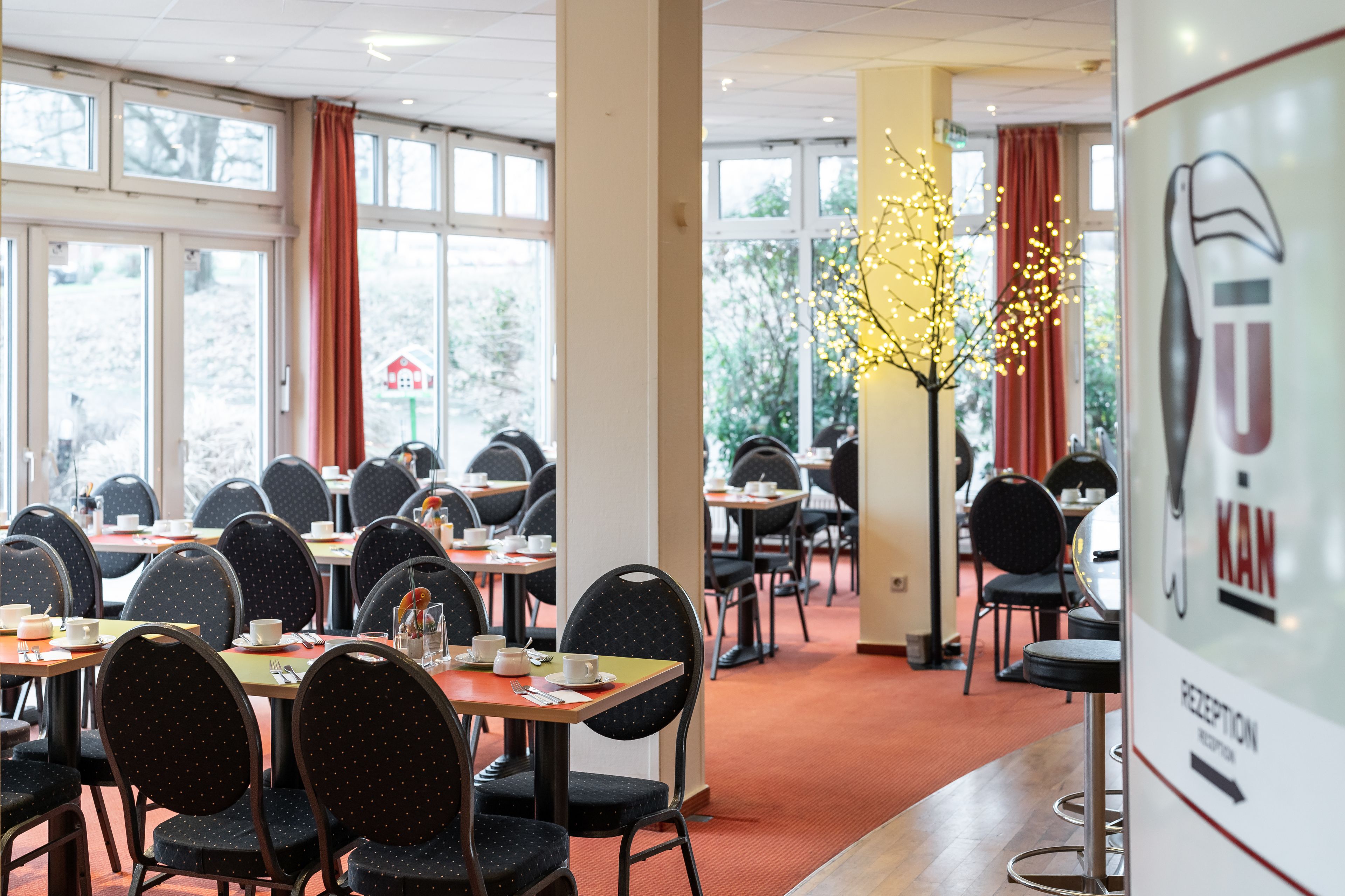A hotel dining area with tables and chairs, a reception desk, and a view of the outdoors.