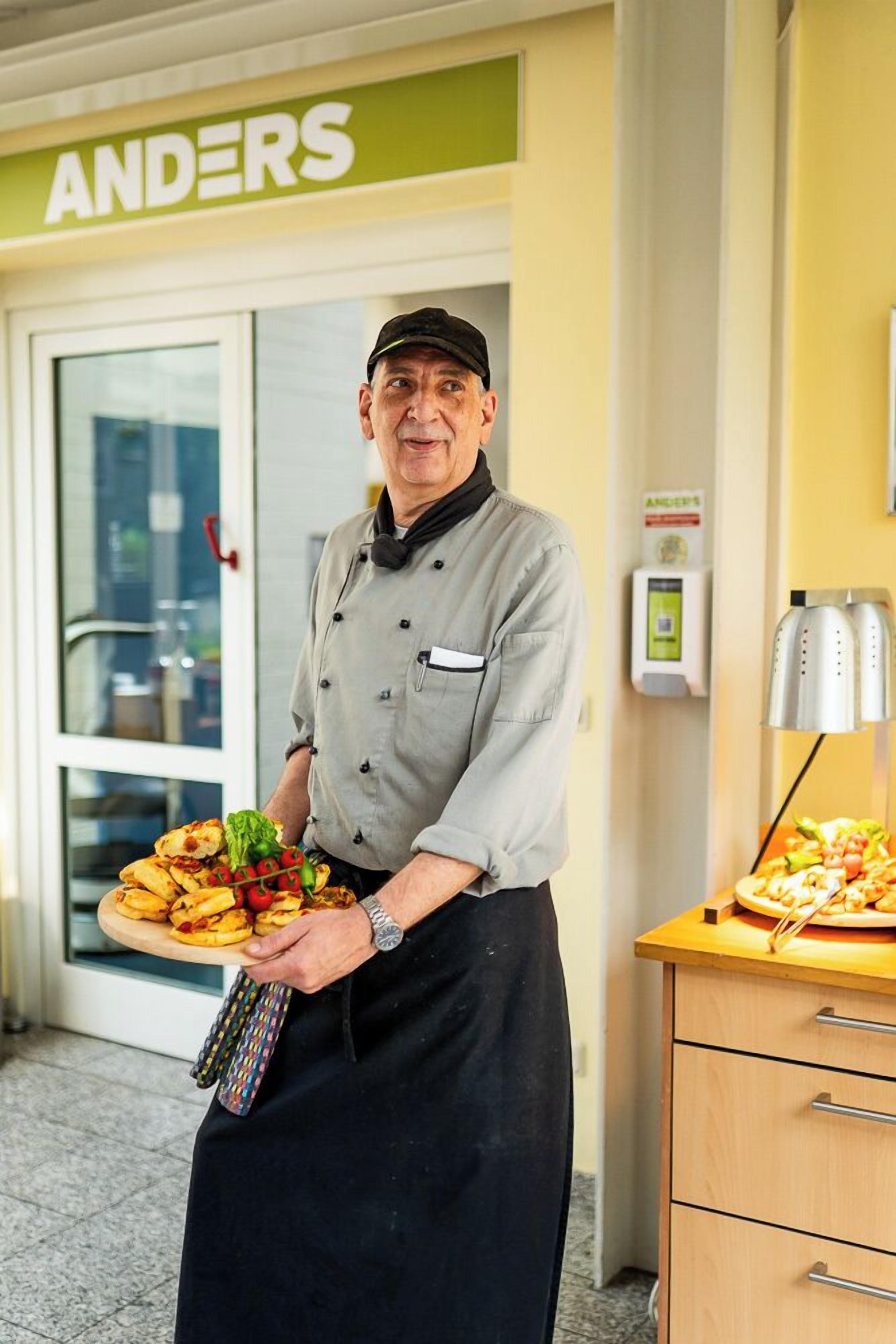 A person in a chef's uniform holding a tray of food in front of a restaurant named 'ANDERS'.