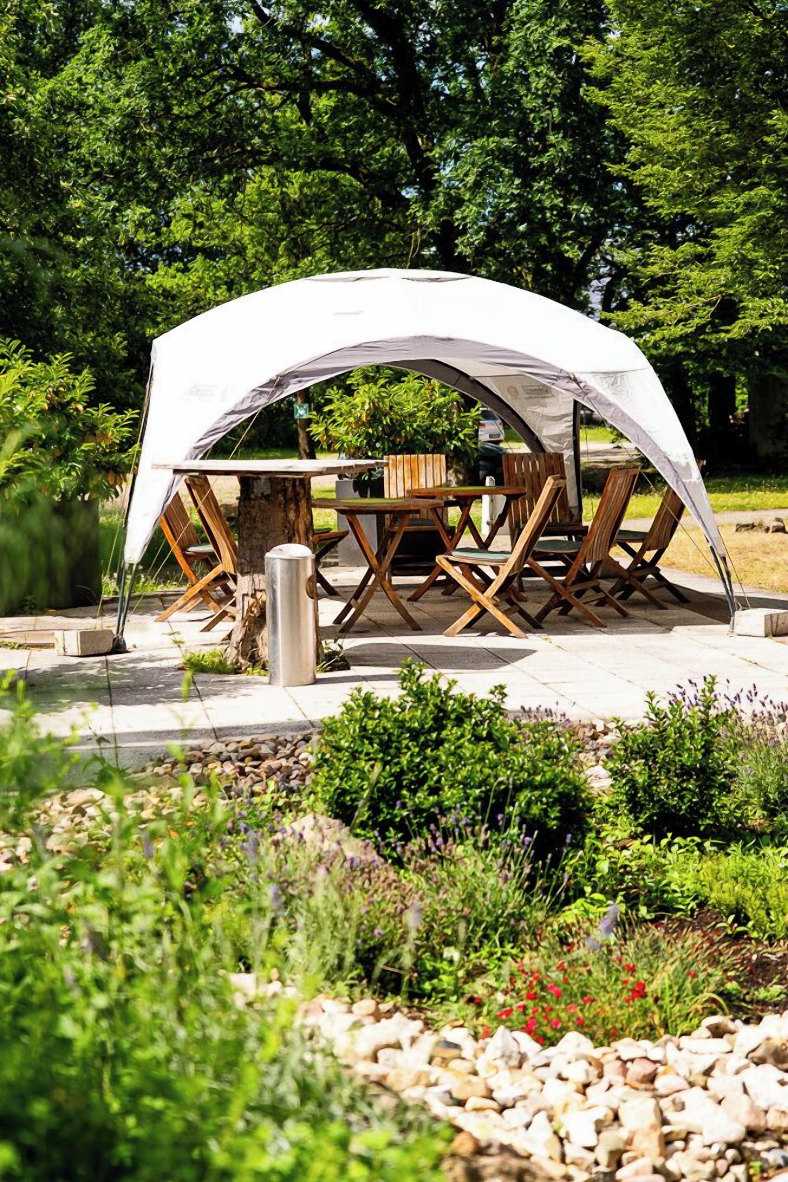 A tent with a table and chairs set up in a garden with trees and plants around.