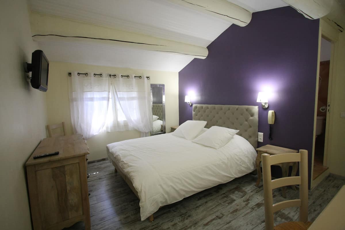 A bedroom with a bed, a wooden nightstand, a chair, a mirror, and a wall-mounted TV.