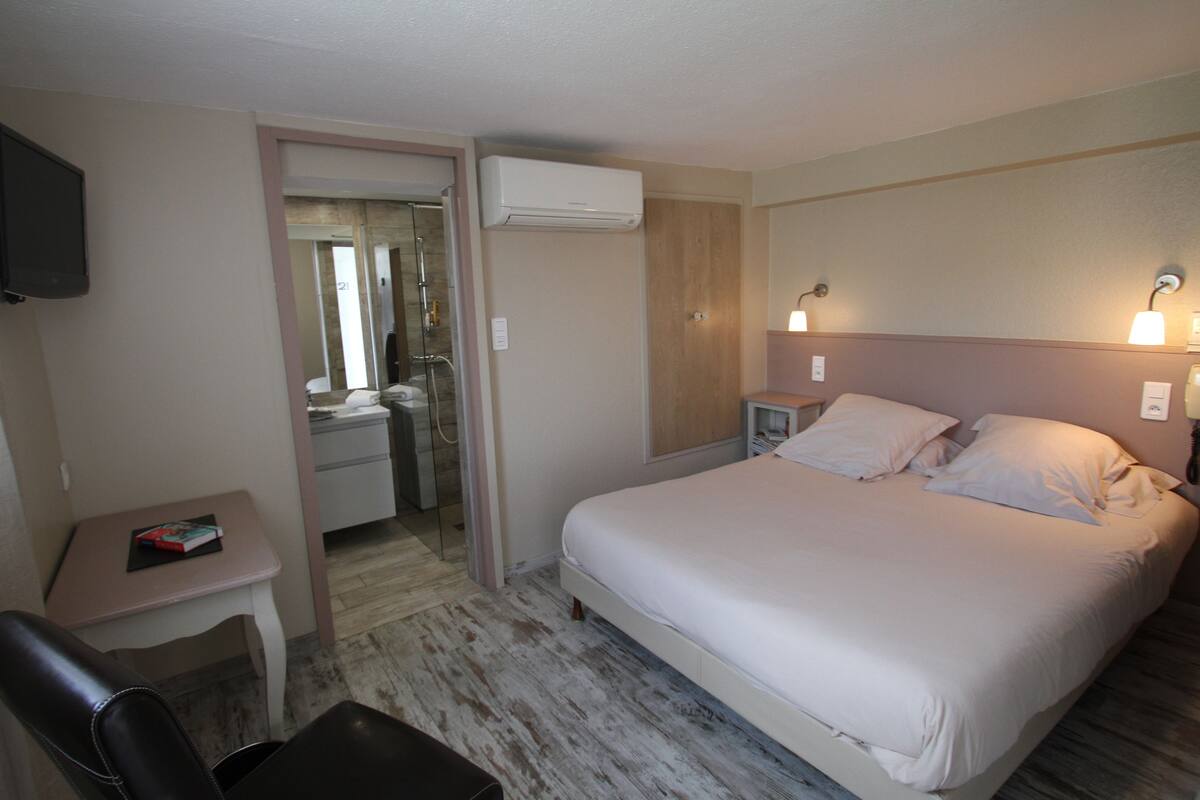 A hotel room with a bed, bedside tables, a desk, a chair, an air conditioning unit, and a television.