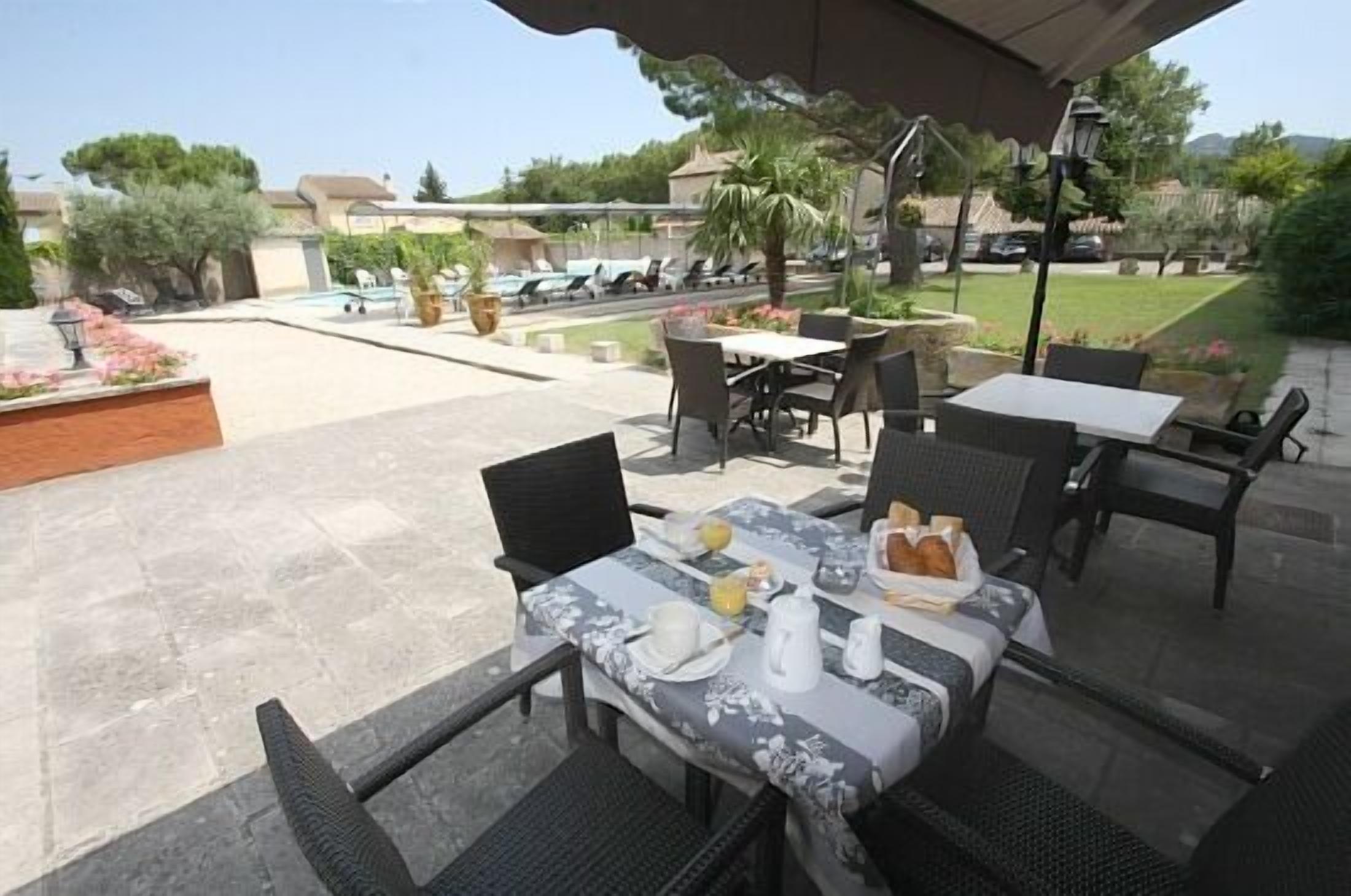Un patio al aire libre con una mesa lista para el desayuno, rodeada de sillas, y una piscina y follaje en el fondo.