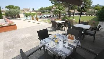 Un patio al aire libre con una mesa lista para el desayuno, rodeada de sillas, y una piscina y follaje en el fondo.