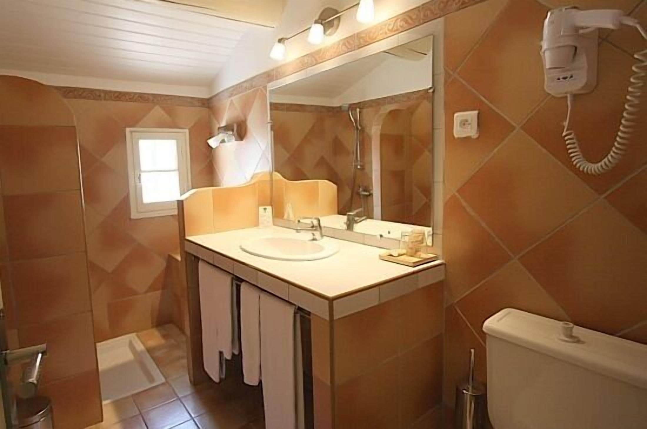 A bathroom with a sink, mirror, and toilet. There is a window and a hairdryer on the wall.