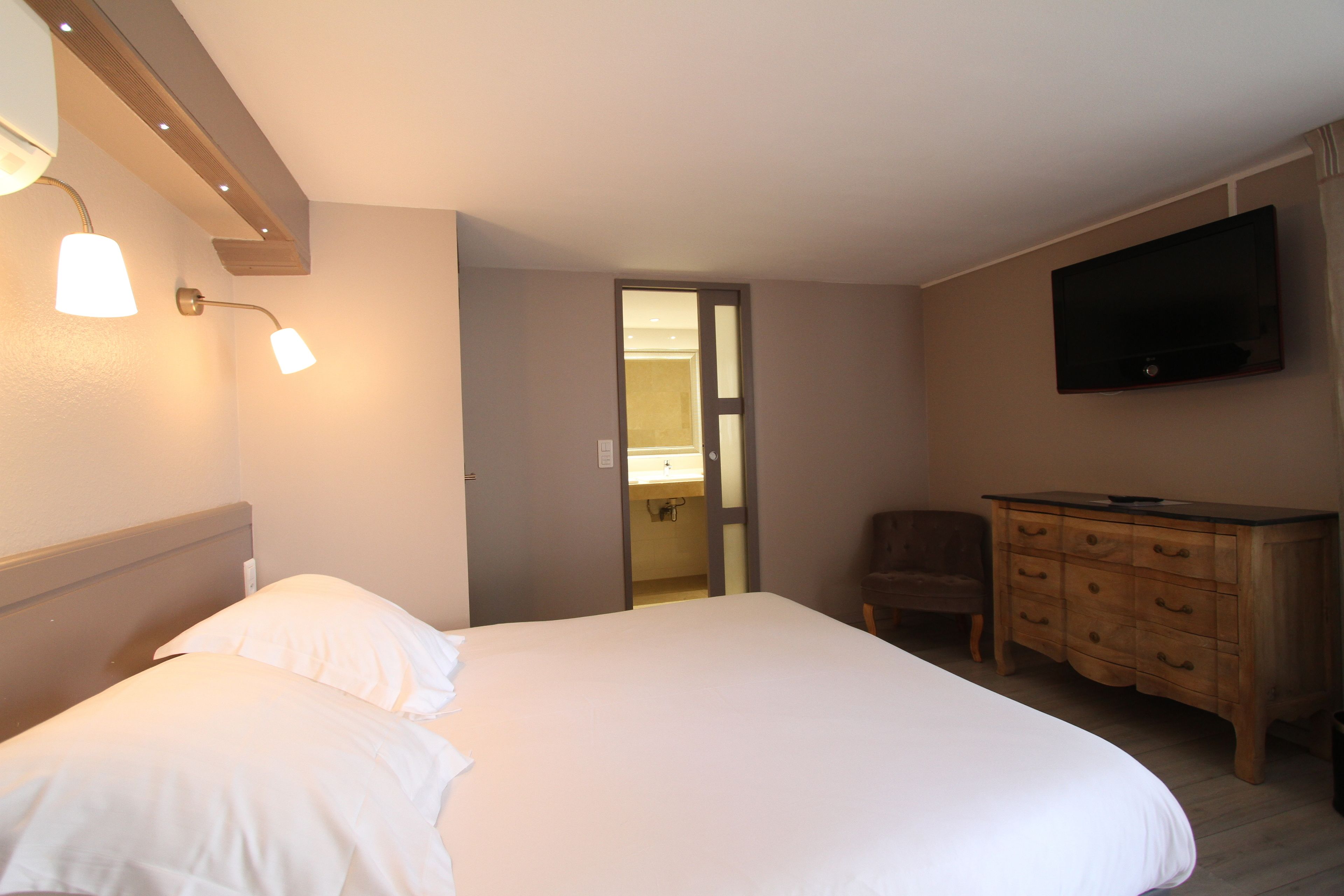 A hotel room with a large bed, a wooden dresser, a television mounted on the wall, and a door leading to a bathroom.