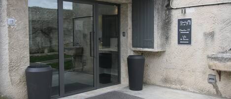 A building entrance with a glass door, two black trash bins, a doormat, and a sign indicating room numbers and a fitness room.