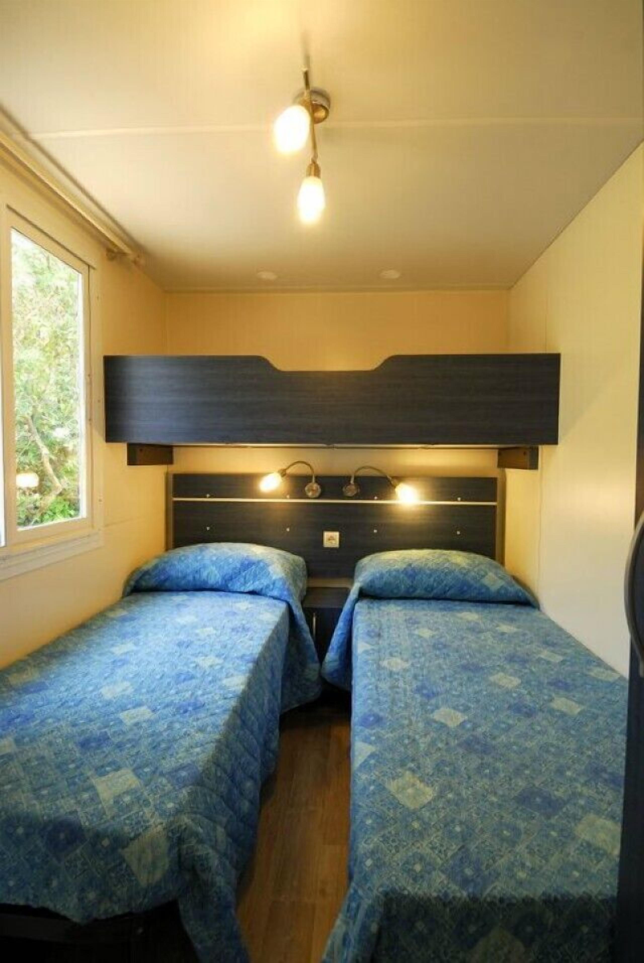 A small room with two beds, each with a patterned blue bedspread. There is a window on the left side, and a ceiling light fixture above the beds.