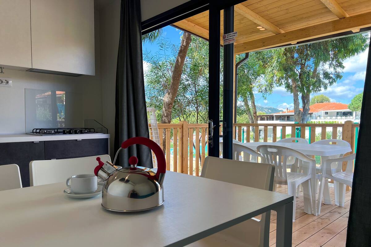 Une cuisine avec une bouilloire sur une table, une tasse, et une vue sur une terrasse extérieure avec des chaises et une rambarde.