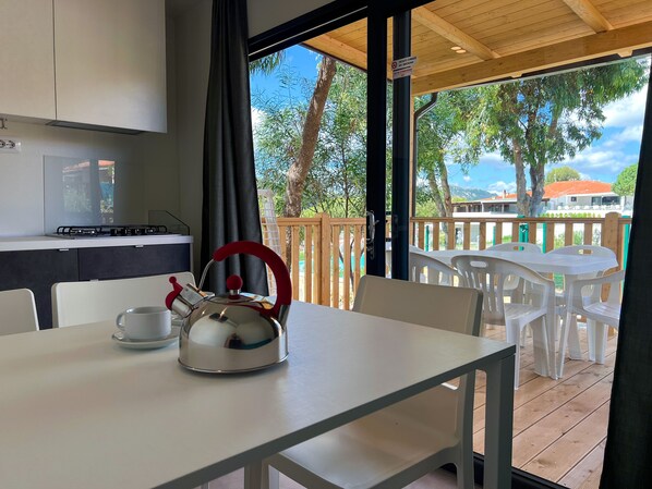 A kitchen with a kettle on a table, a cup, and a view of an outdoor deck with chairs and a railing. - Camping Village Capo D'Orso (Palau)