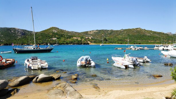 A marina with various boats docked, clear water, and a hilly landscape in the background. - Camping Village Capo D'Orso (Palau)