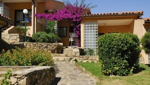 A building with a stone pathway leading to a door, surrounded by greenery and a purple flowering plant. - Camping Village Capo D'Orso (Palau)