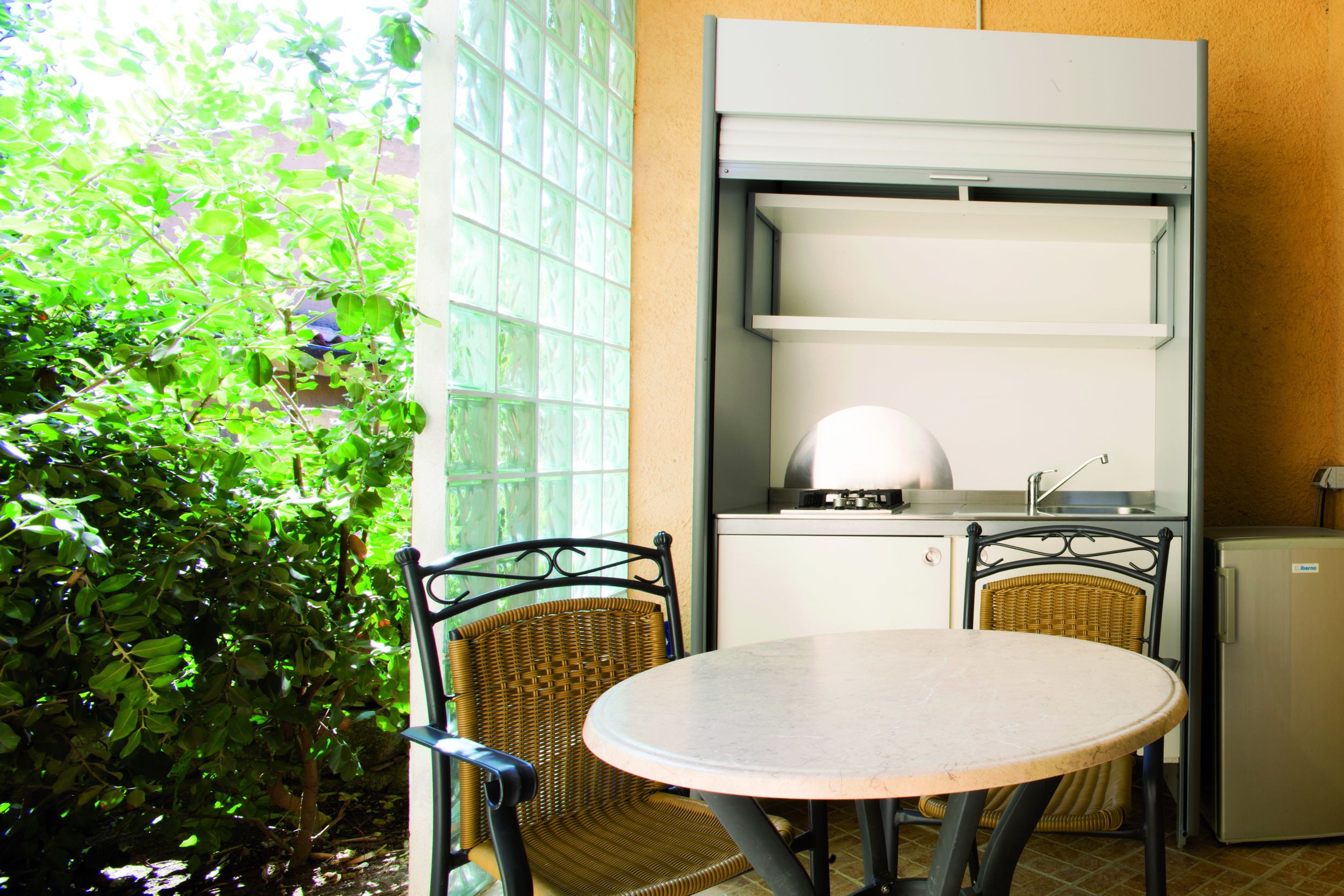 A compact kitchen with a built-in oven, a small refrigerator, and a dining area with two wicker chairs and a round table.
