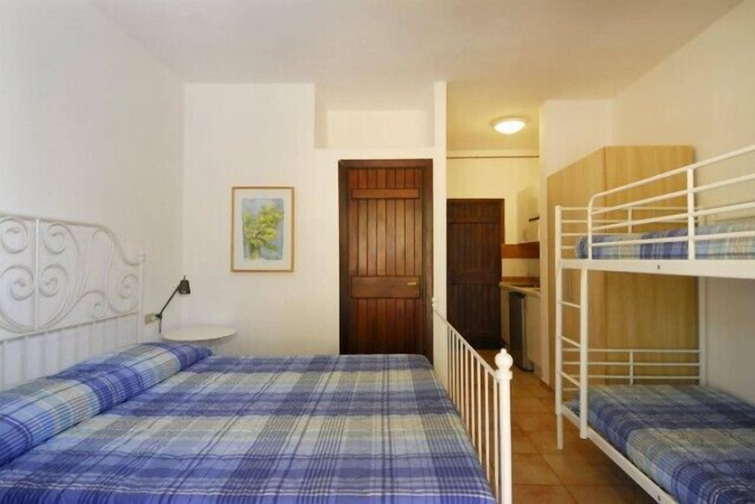 Una habitación con dos camas, una con cabecera y otra tipo litera. Hay una mesita con una lámpara, una puerta y un cuadro enmarcado en la pared.