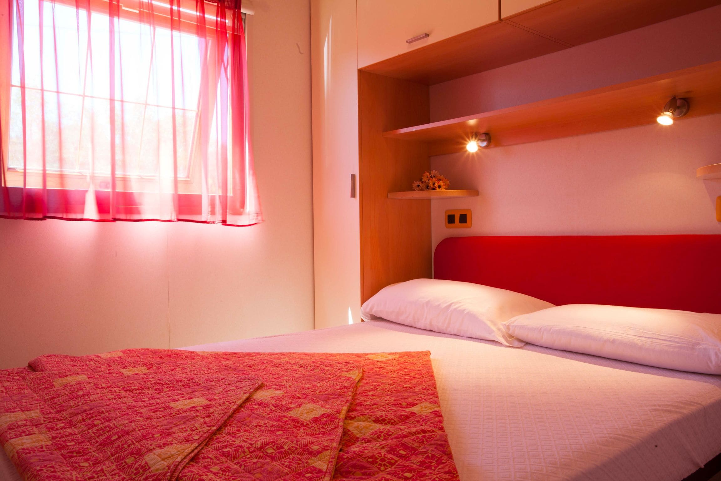 Una habitación de hotel con una cama, una ventana con cortinas traslúcidas, un estante con adornos y un cabecero rojo.