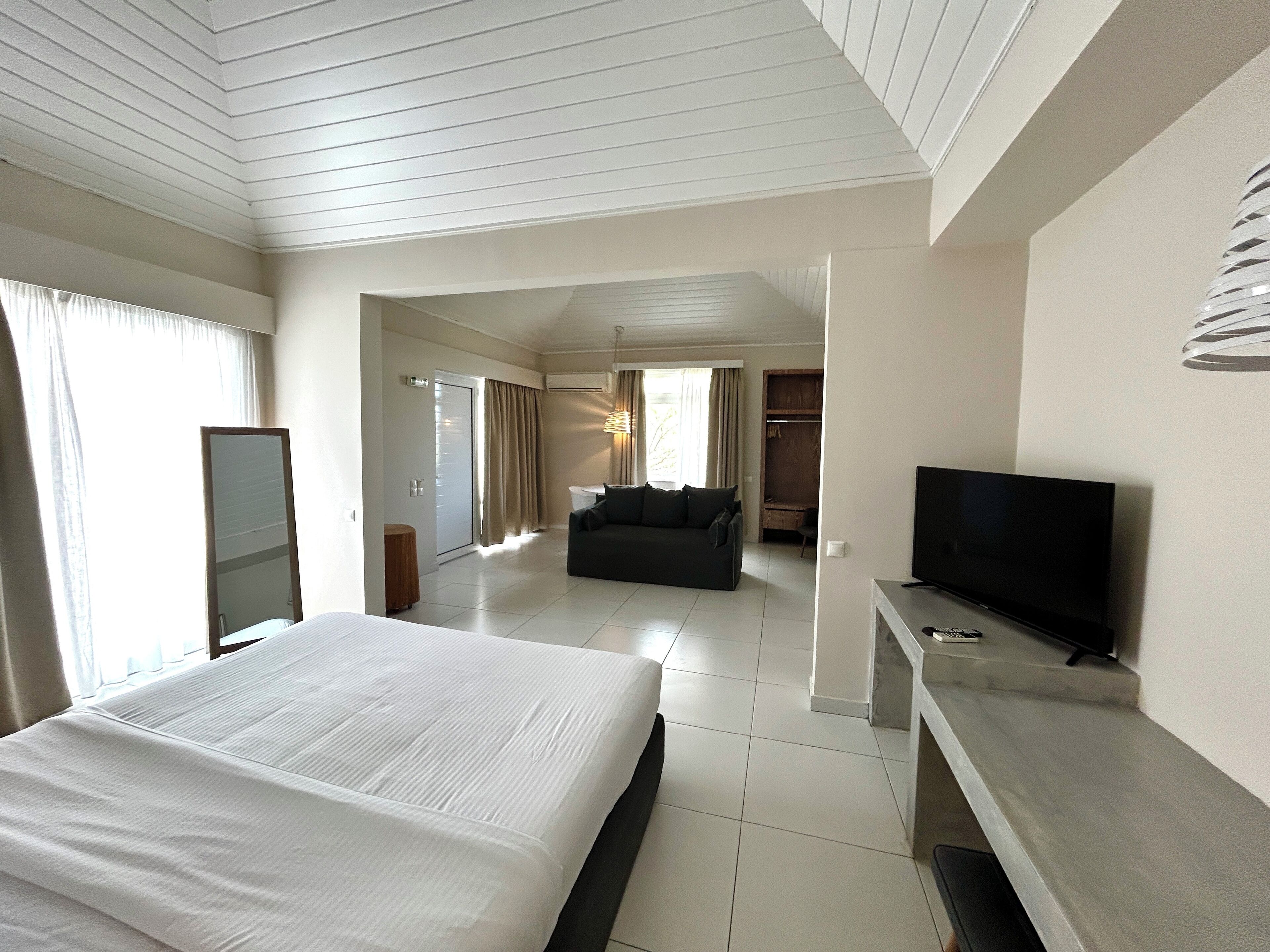 Habitación de hotel con una cama grande, televisión, sofá y escritorio. Se observa un armario y una puerta que conduce a otra habitación.