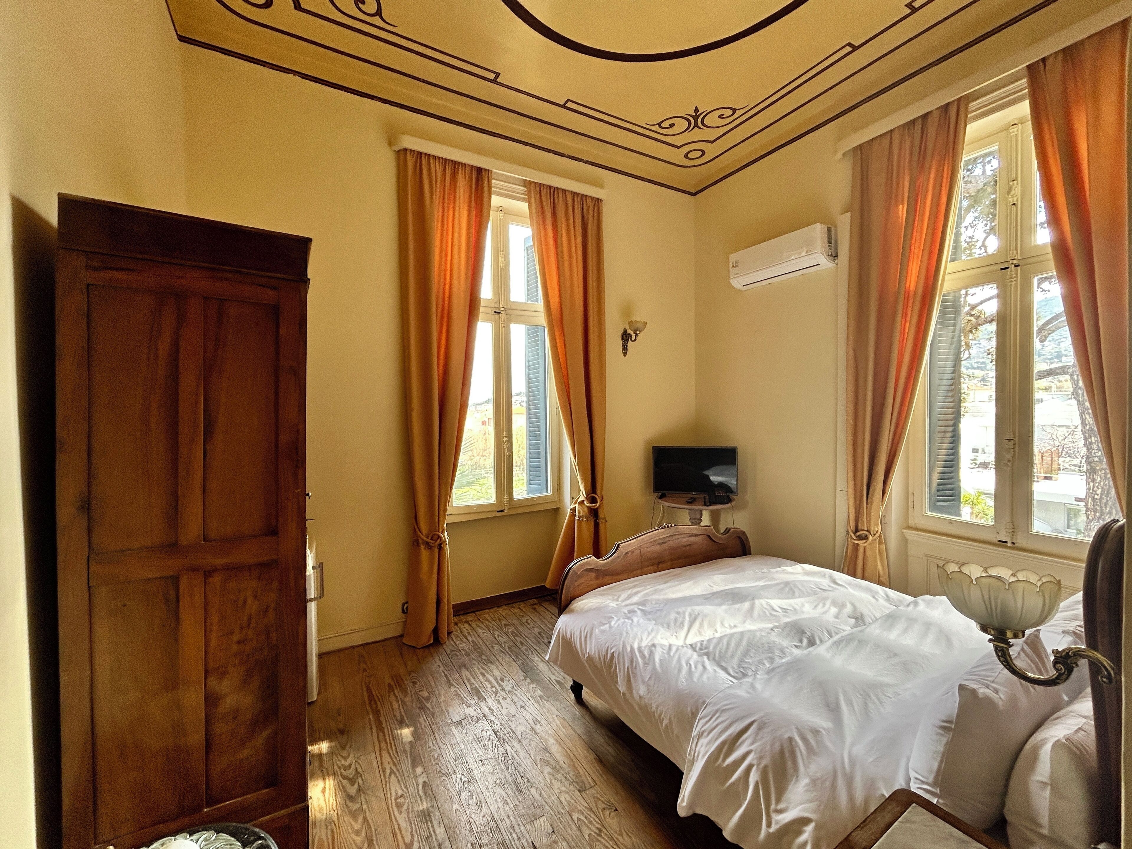 A bedroom with a wooden wardrobe, a bed with white bedding, an air conditioning unit, and a television on a small table.